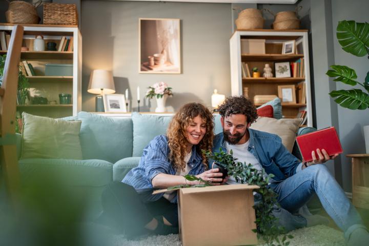 Happy couple moving into a new apartment, sitting on the floor in the living room while unpacking a cardboard box with a houseplant, laughing and enjoying the process